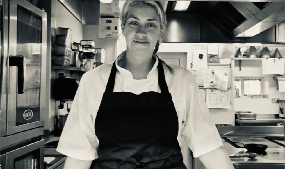 Chef standing in a commercial kitchen, wearing a black apron and white jacket, smiling at the counter