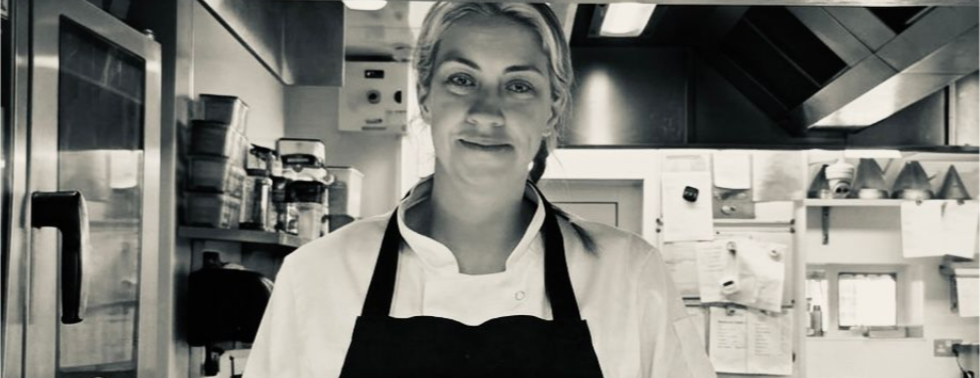 Chef standing in a commercial kitchen, wearing a black apron and white jacket, smiling at the counter