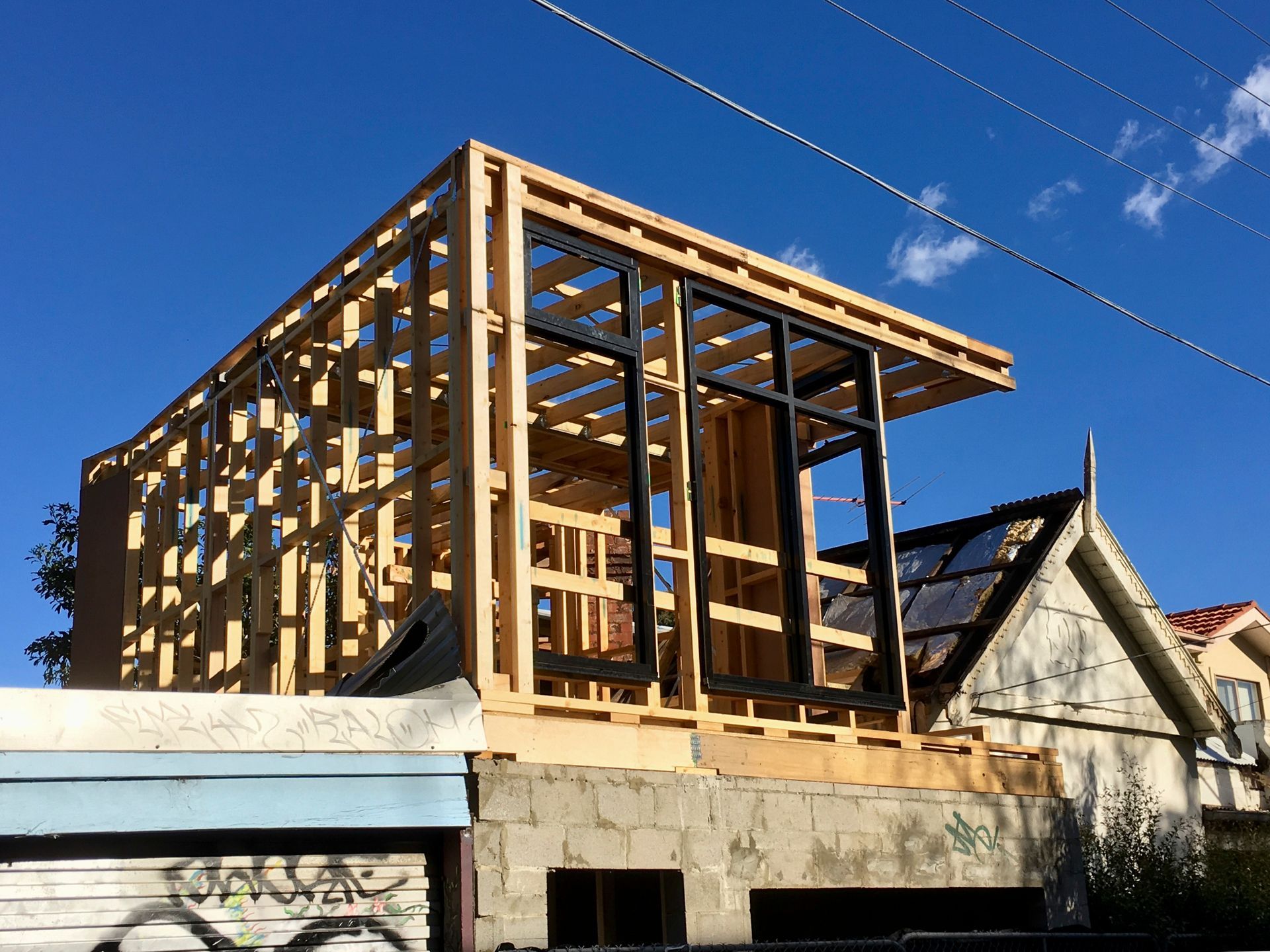 Extension à ossature bois en construction sur un bâtiment, avec de grandes fenêtres à encadrement noir. Ciel bleu dégagé.