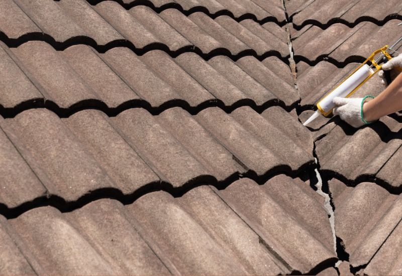 Person in gloves sealing crack between brown roof tiles with caulk gun.