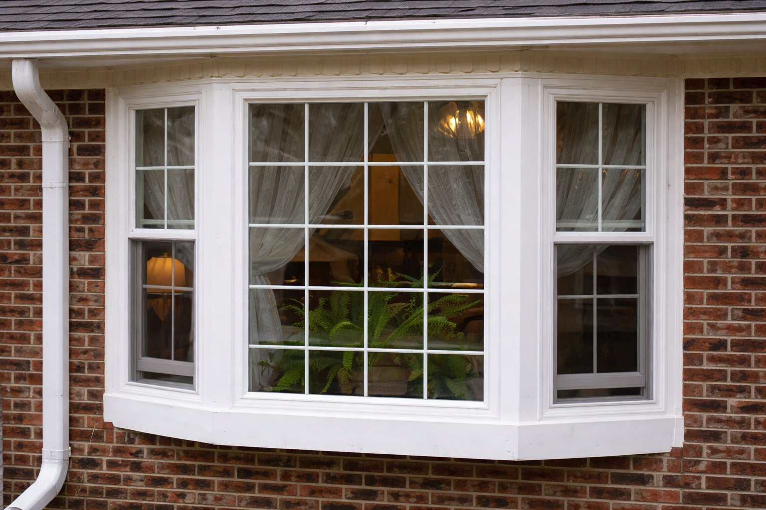 Bay window with white frame and grid pattern, set in brick wall, with plants and light visible through glass.