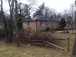 Roofers working on a brick house roof with trees in the foreground.