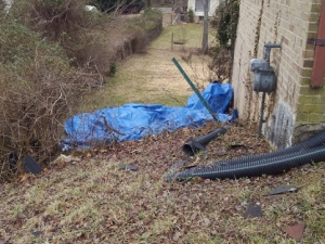 Blue tarp covering debris near a brick building with a gas meter and drainage pipes.