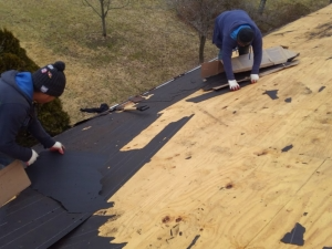 Two people working on a roof, installing a black underlayment. Wooden boards and bare trees are in the background.