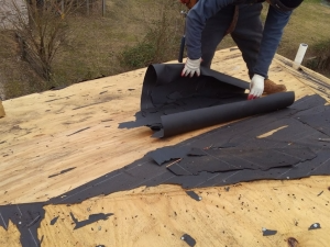 Person unrolling black roofing underlayment on wooden rooftop.