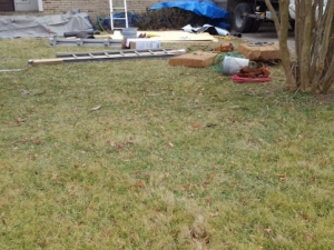 Lawn with construction materials including a ladder, covered by a partially visible blue tarp in front of a brick building.