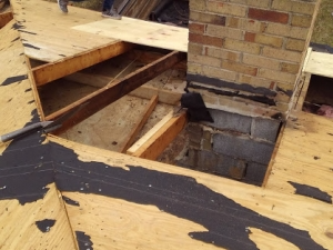 Rooftop with plywood partially removed, exposing framing around a brick chimney, sections of roofing felt visible.