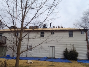 Workers on a roof removing old shingles from a two-story white house. Blue tarps protect the ground.