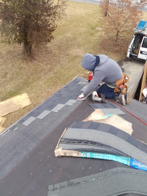 Roofer kneeling on a roof, installing shingles, wearing a gray hoodie and tool belt, daylight.