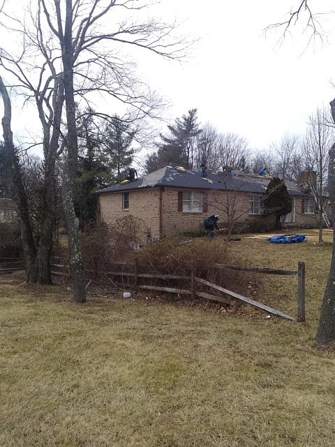 A brick house with a damaged roof, trees, and a wooden fence. Overcast day.