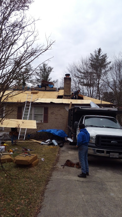 Roofers working on a house with a dump truck parked in the driveway. A person stands nearby. Cloudy day.