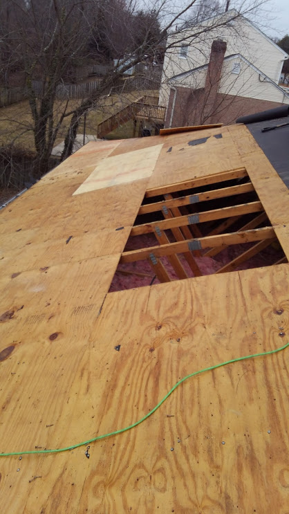 Rooftop construction: Plywood sheathing installed, opening for access, insulation visible, green cable, and distant houses.
