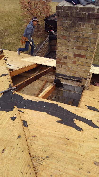 Roofer near a chimney on a partially torn-up roof. The roofer is wearing a tool belt and holding a tool.