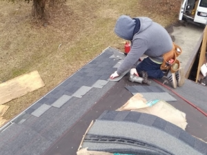 Roofer in a gray hoodie kneels on a dark shingle roof, installing shingles with a nail gun.