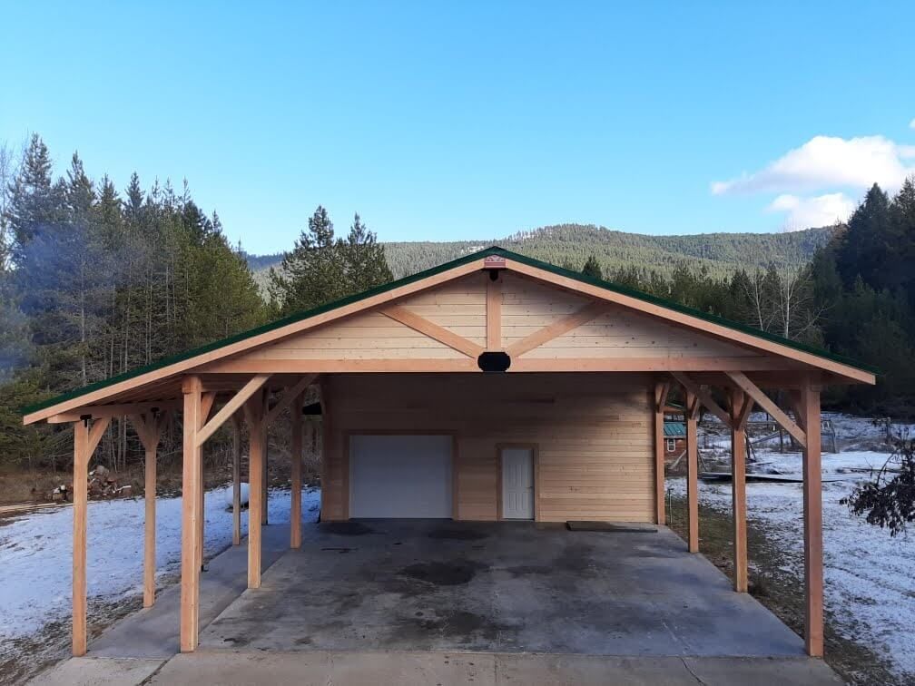 A wooden garage with a green roof is surrounded by snow and trees.