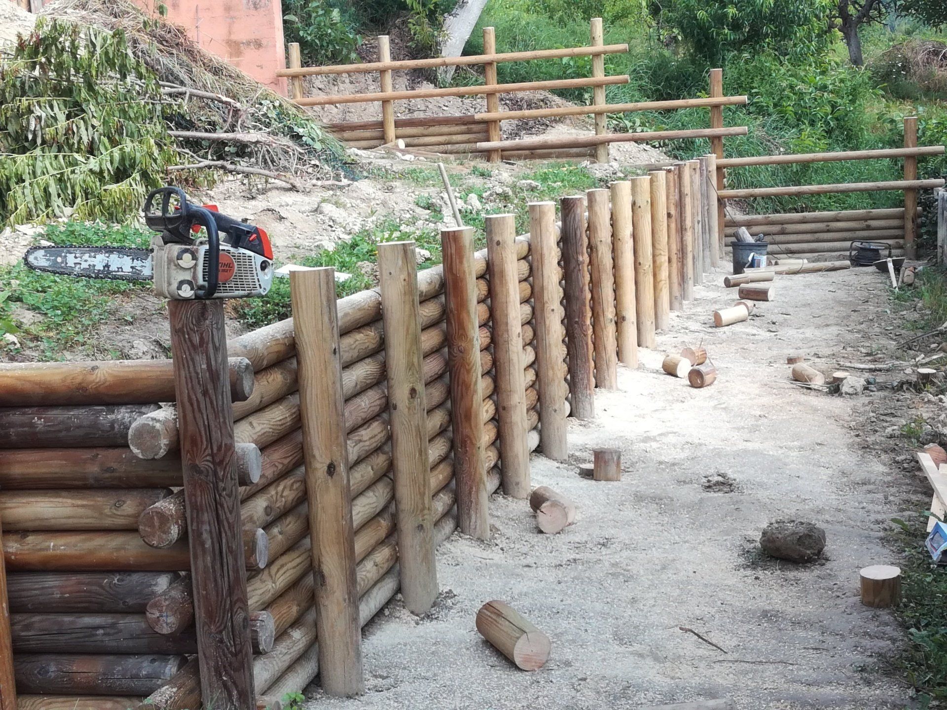 Staccionata in legno in costruzione in un giardino; una motosega è appoggiata su un palo.