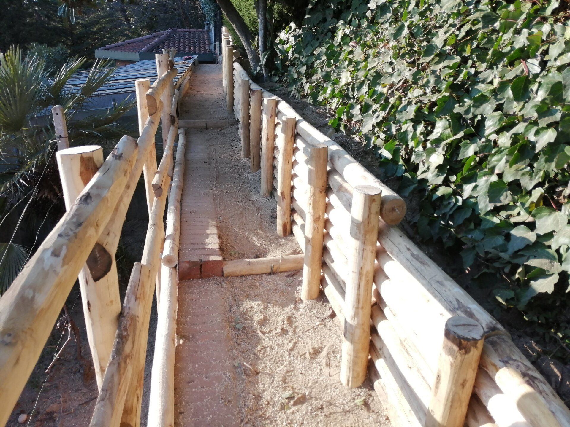 Passerella in legno con ringhiere in tronchi e sentiero di ghiaia, accanto a un muro di edera.