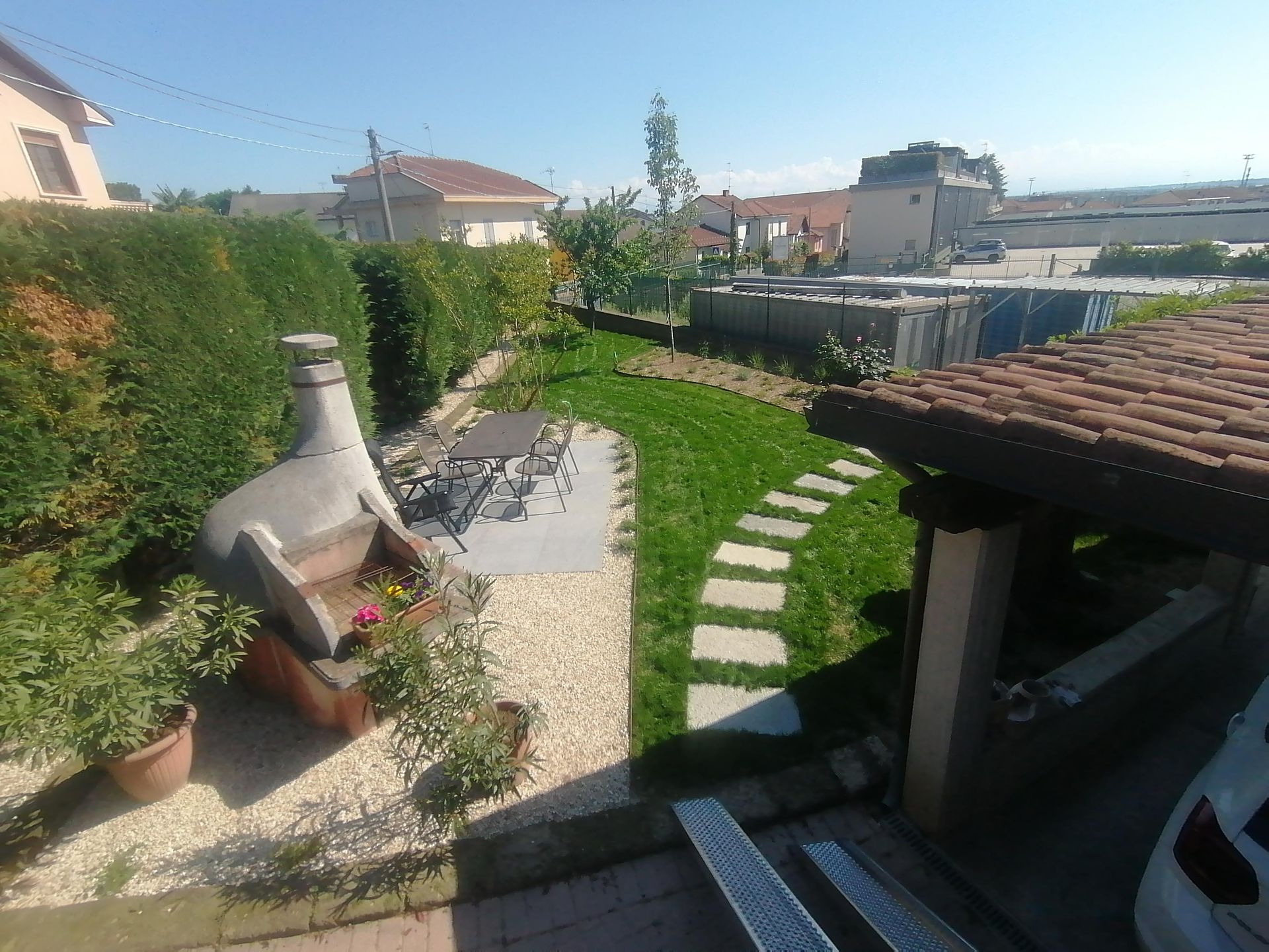 Un cortile con barbecue, pietre per camminare e verde sotto un cielo soleggiato.