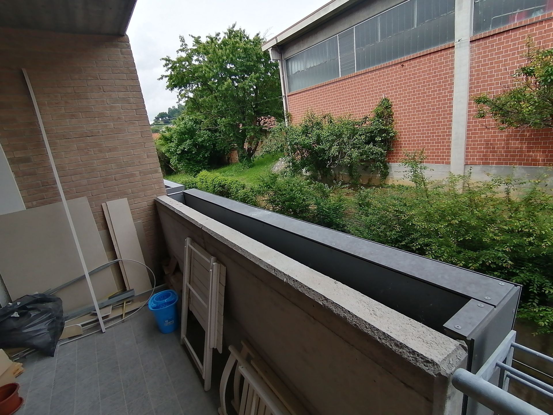 Balcone con lunga fioriera, edificio sullo sfondo, oggetti vari contro il muro.