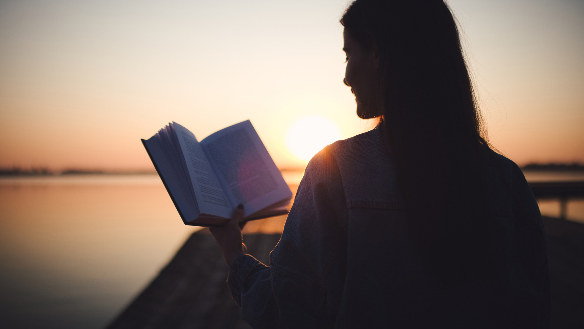 A woman is reading a book by the water at sunset.