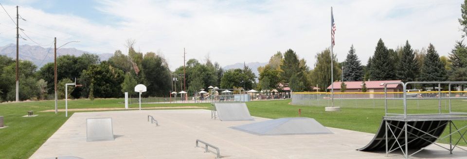 Skatepark with ramps, rails, and a basketball hoop. Trees and mountains in the background. Green grass and a cloudy sky.