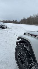 A white Jeep Rubicon in a snowy field, connected by a tow cable to a dark vehicle stuck in the snow ahead.