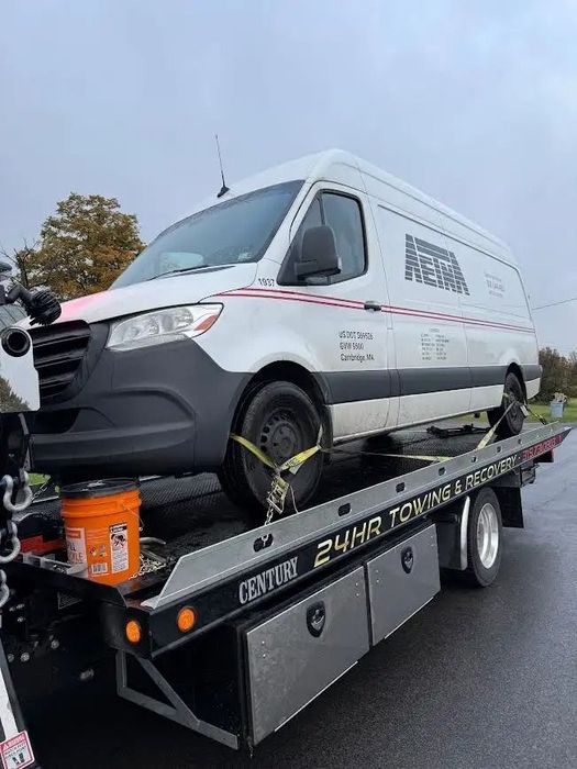 A white delivery van secured with yellow straps on the back of a flatbed tow truck on a cloudy day.