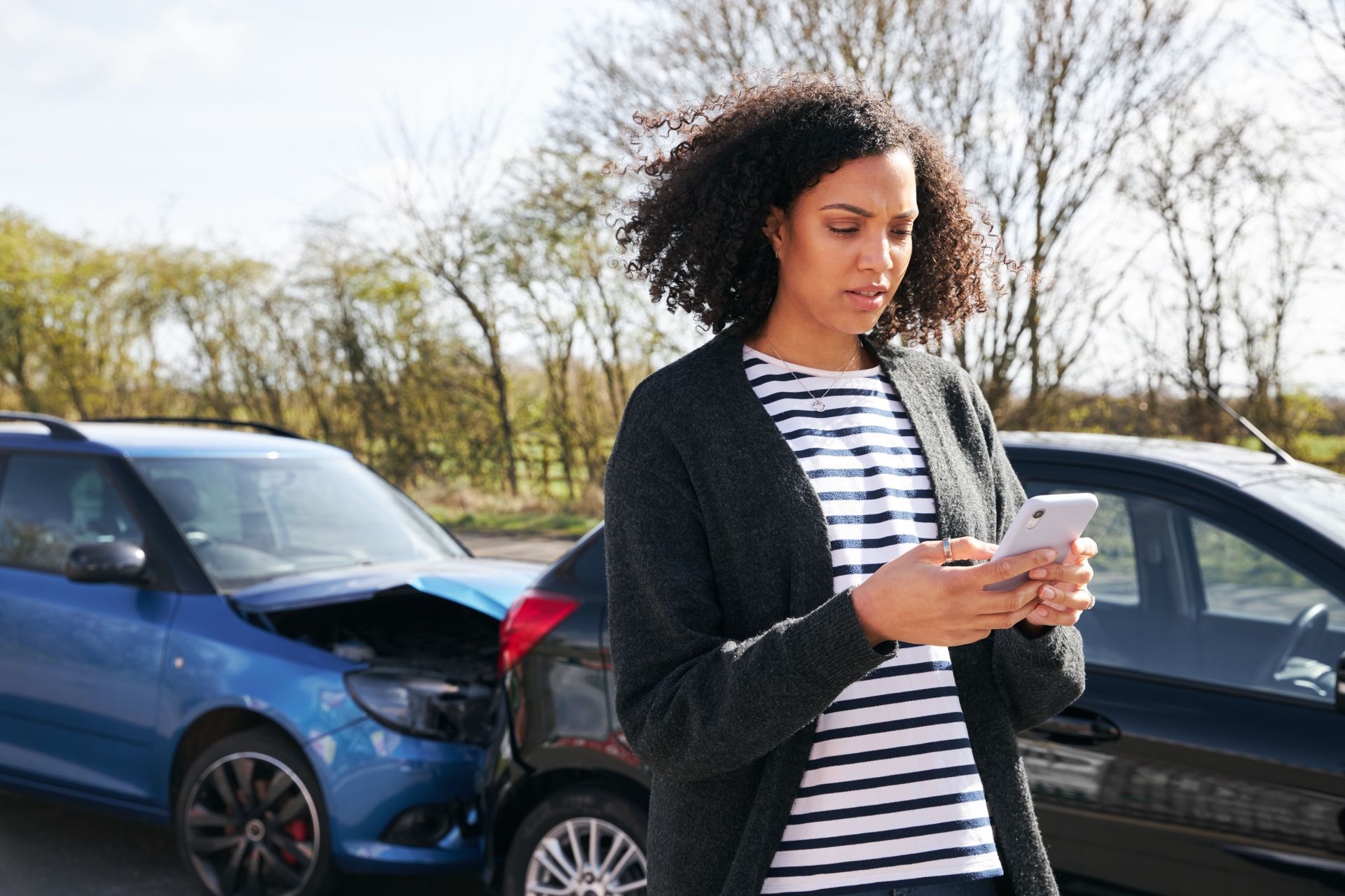 A woman is standing in front of a broken down car looking at her phone.