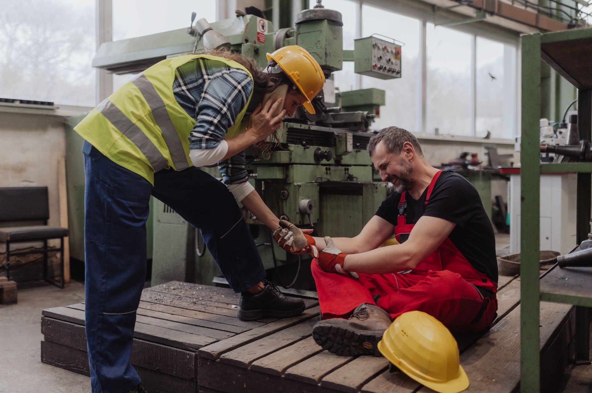 A man is sitting on the ground in a factory while another man helps him.