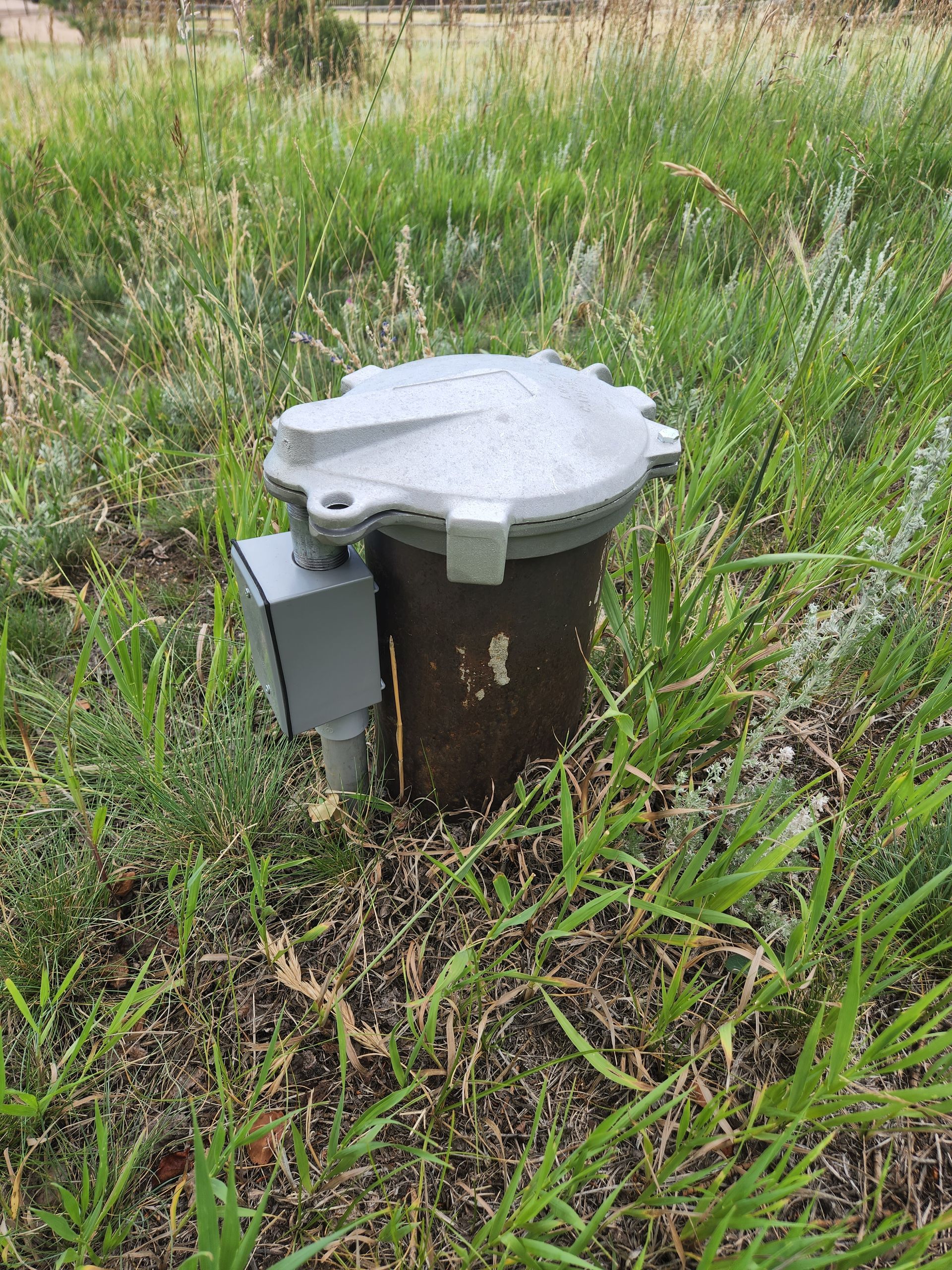 A gray metal well with a lid and electrical box, surrounded by grass.