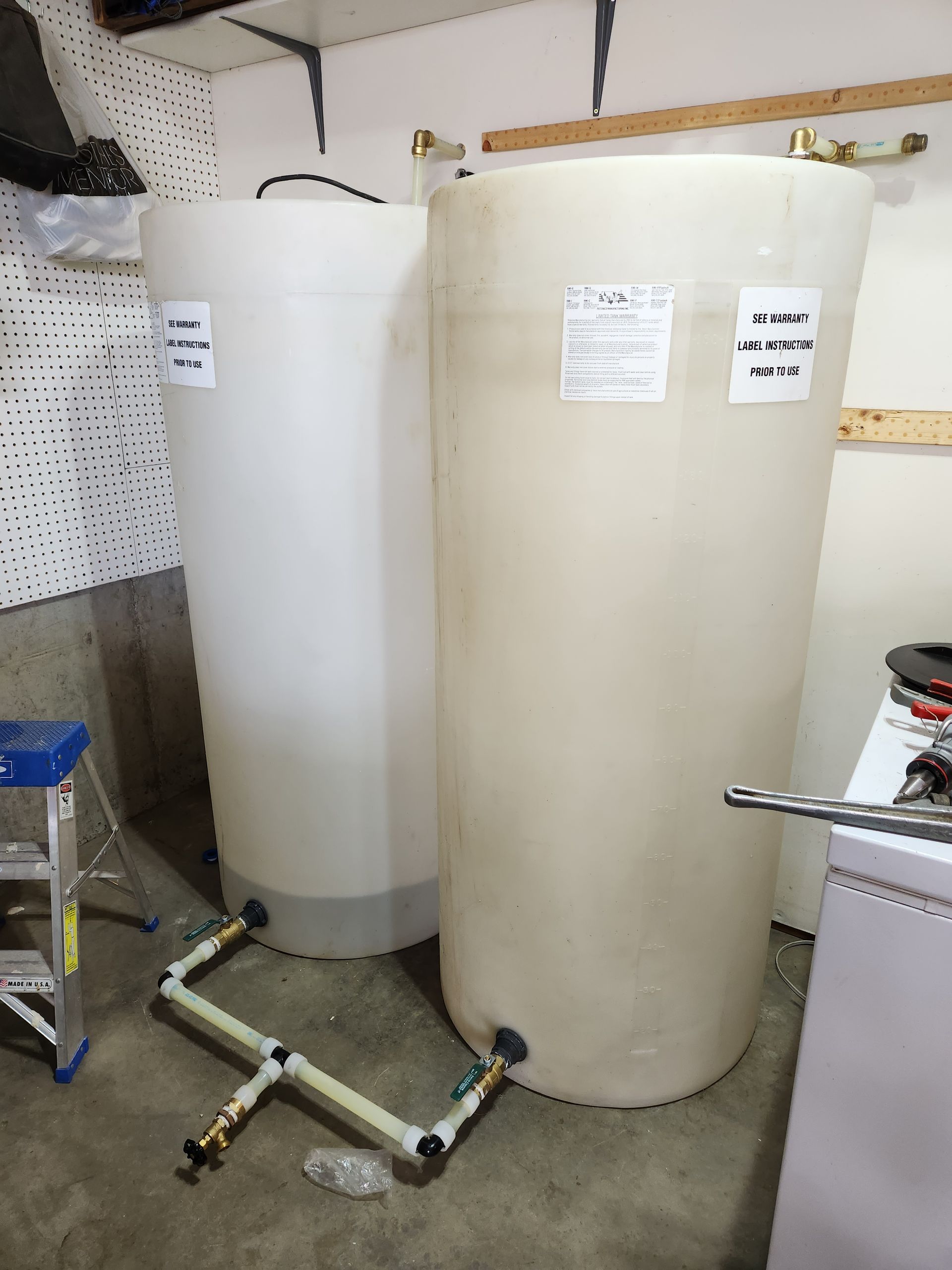 Two large, white plastic water tanks in a basement. Pipes and ladder are visible.