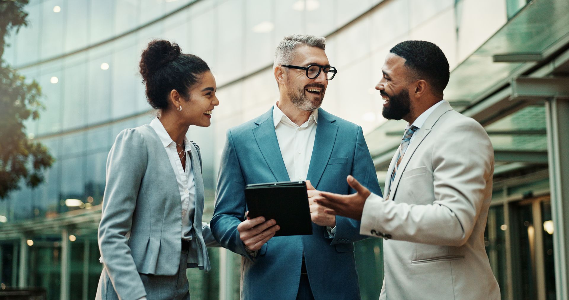 Three people in suits talking outdoors by a building, one holding a tablet.