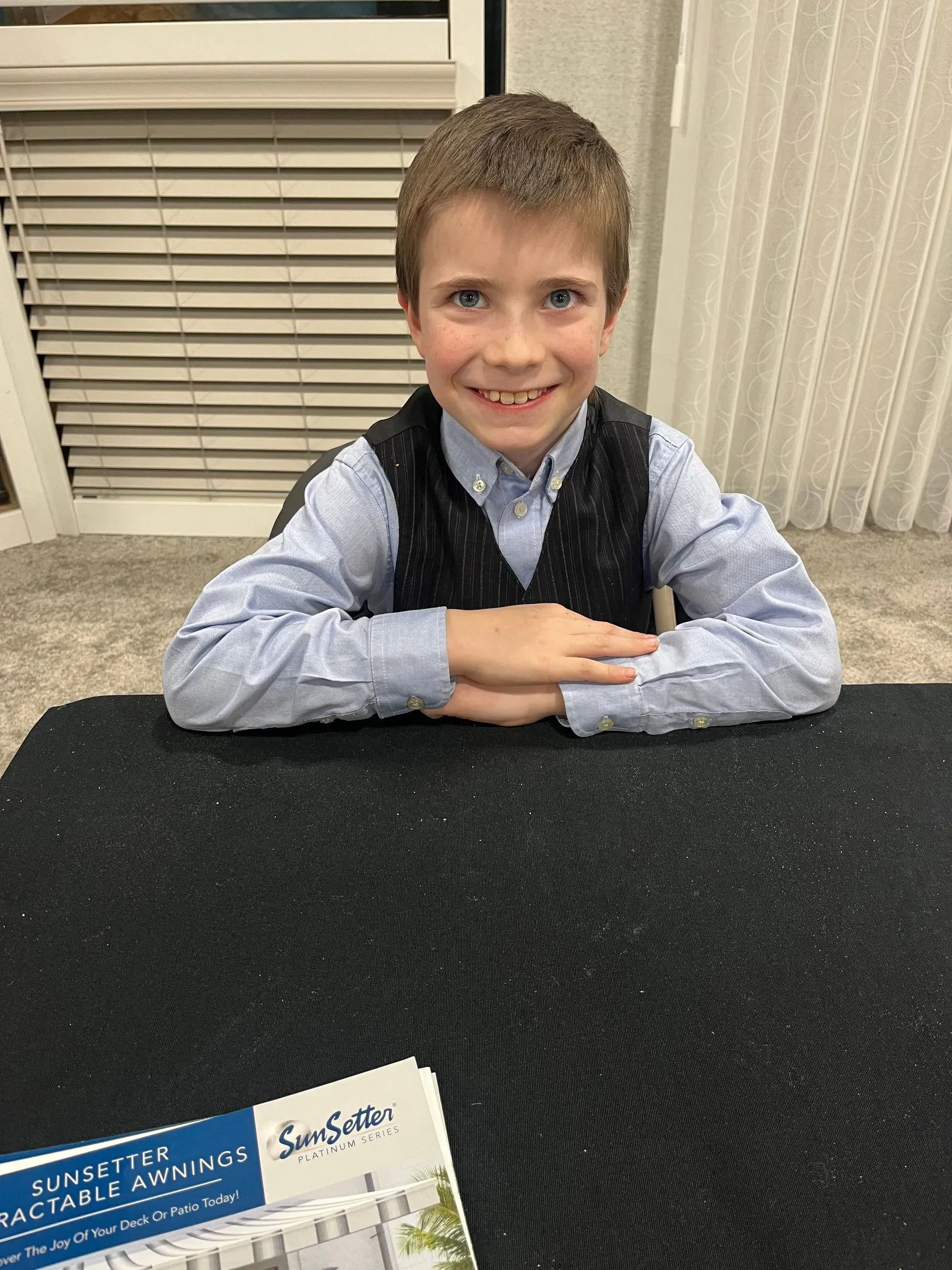 A young boy is sitting at a table with his hands folded and smiling.
