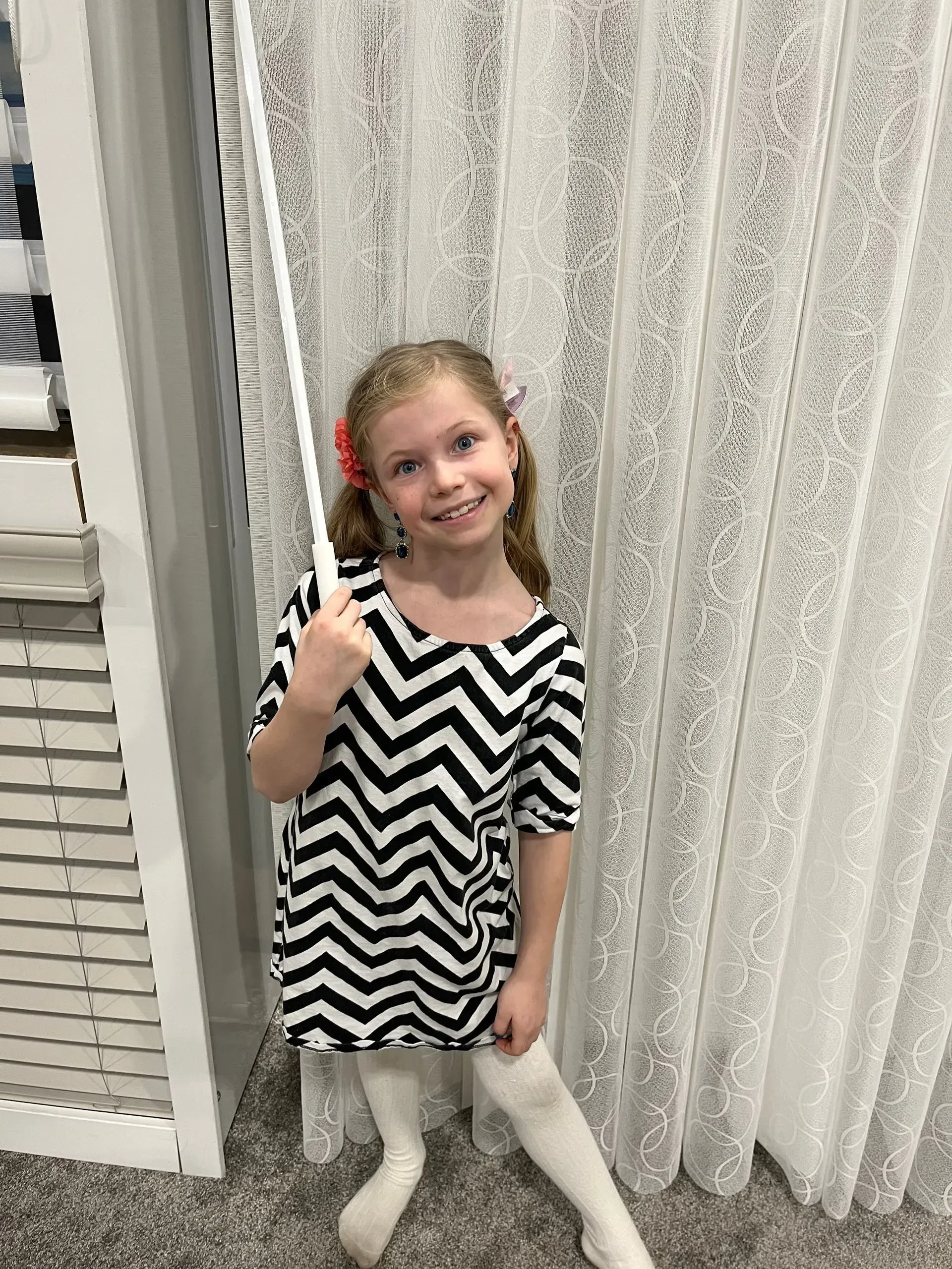 A little girl in a black and white striped dress is standing in front of a white curtain.