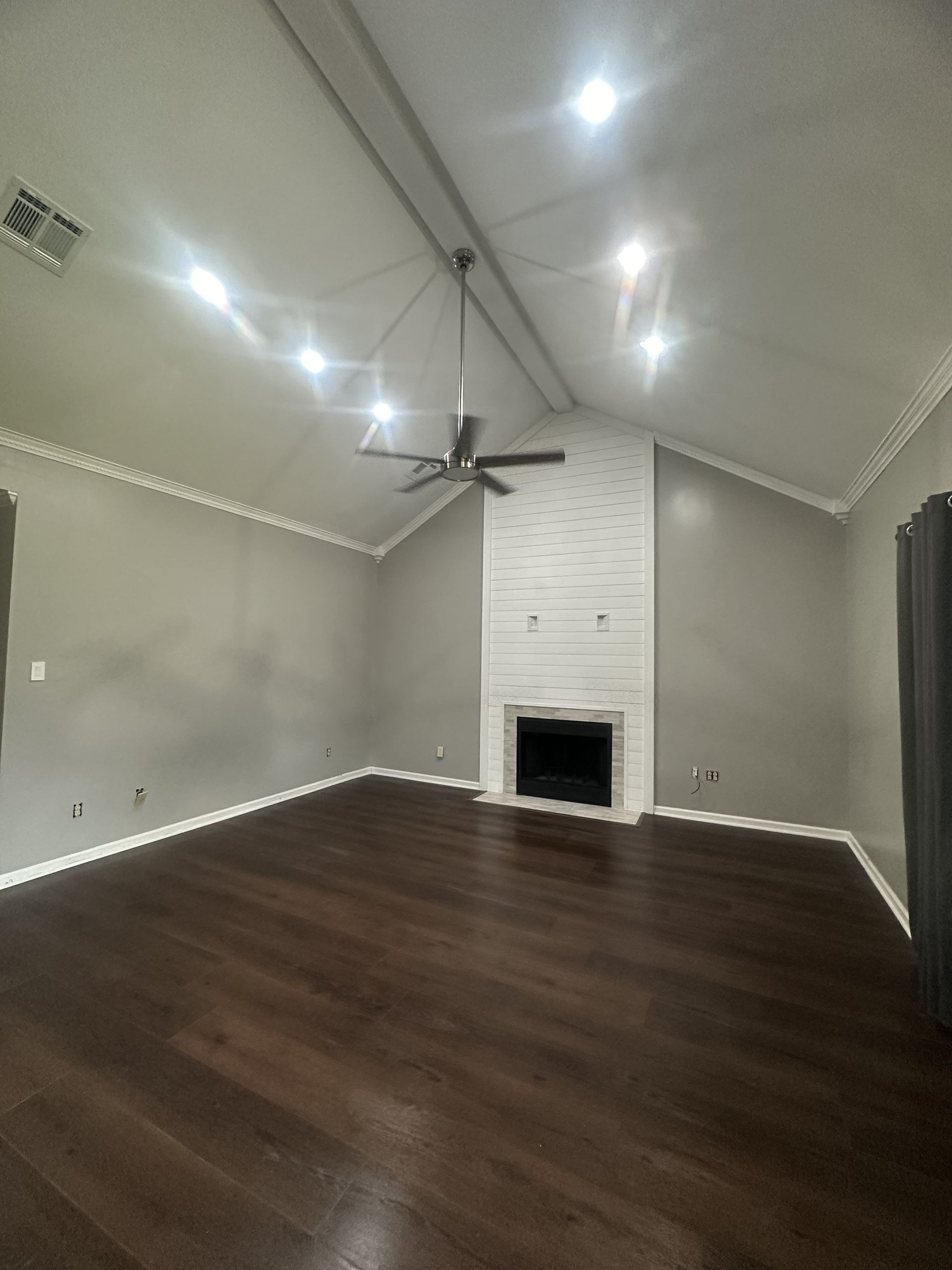 Empty living room with wood floor, white fireplace, grey walls, and ceiling fan.
