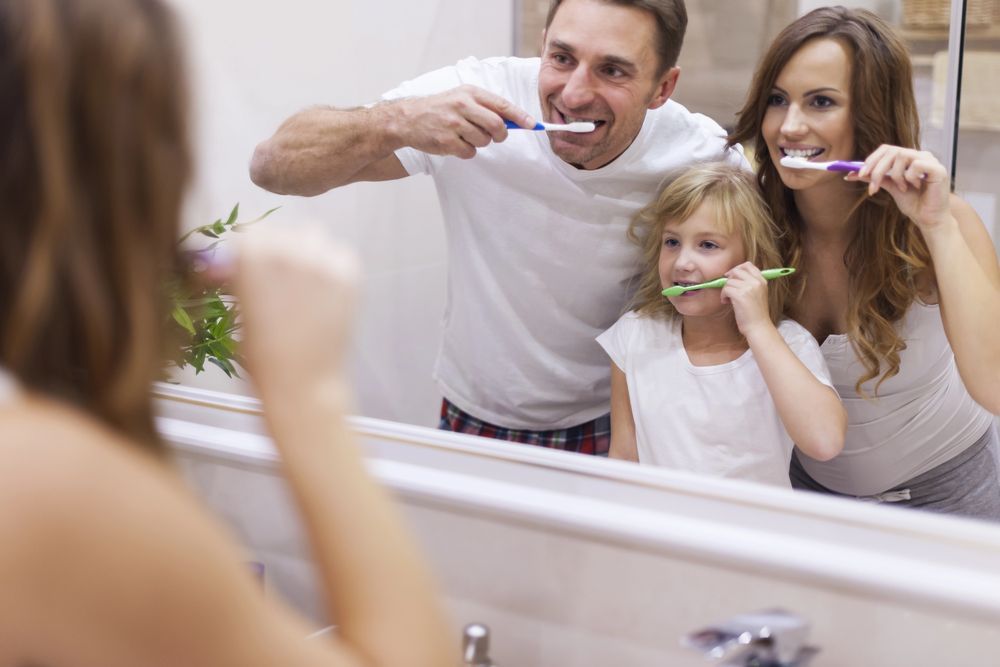 Una famiglia si lava i denti insieme in un bagno, riflessa in uno specchio.