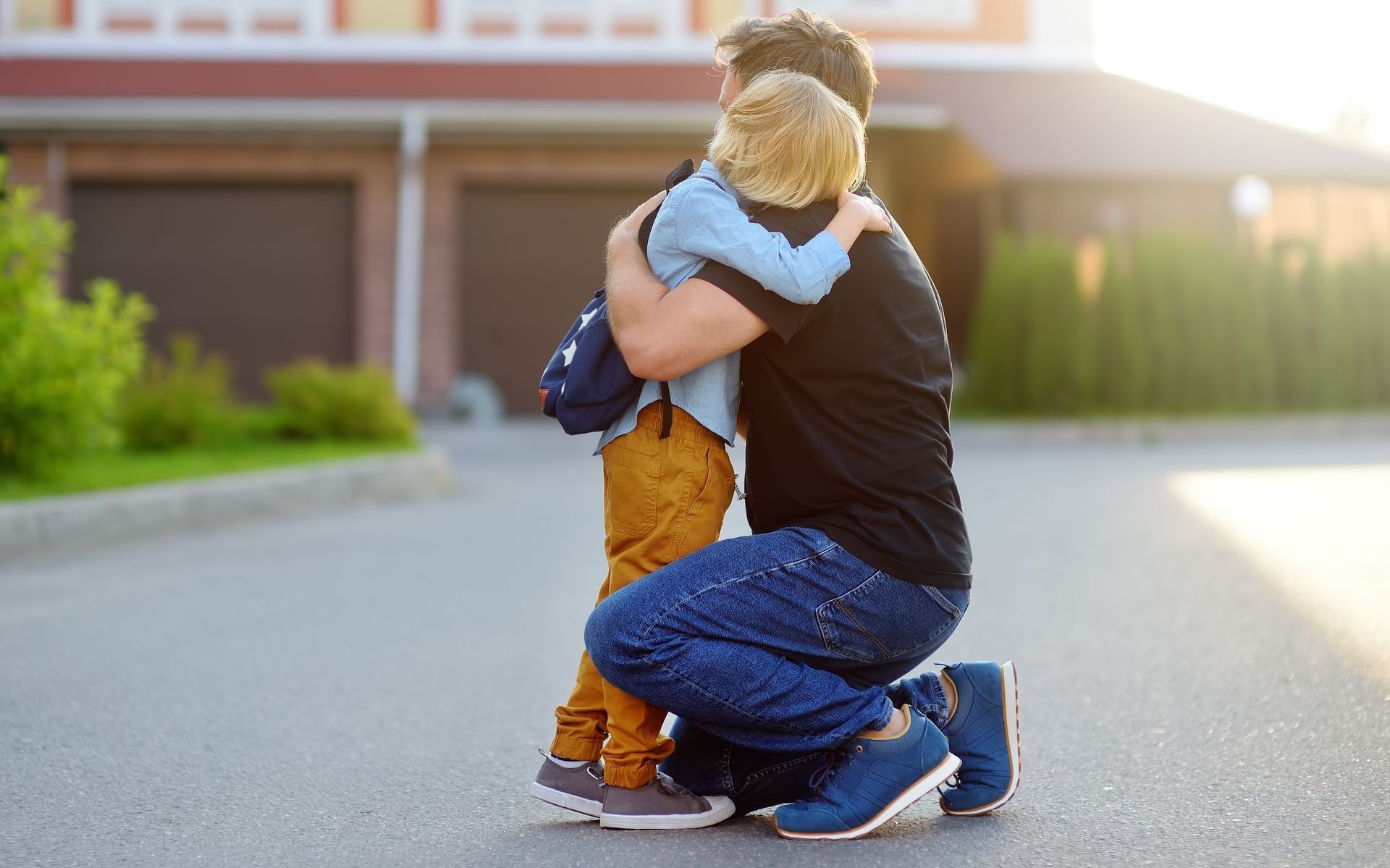 A man is kneeling down and hugging a little boy with a backpack.