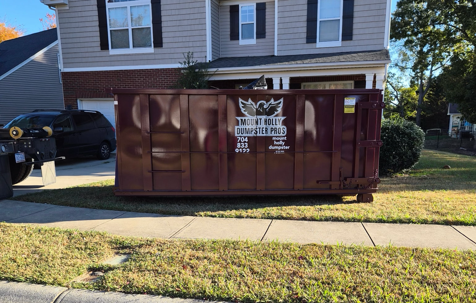 Maroon dumpster on grass in front of a gray house.  A black vehicle is parked on the left.