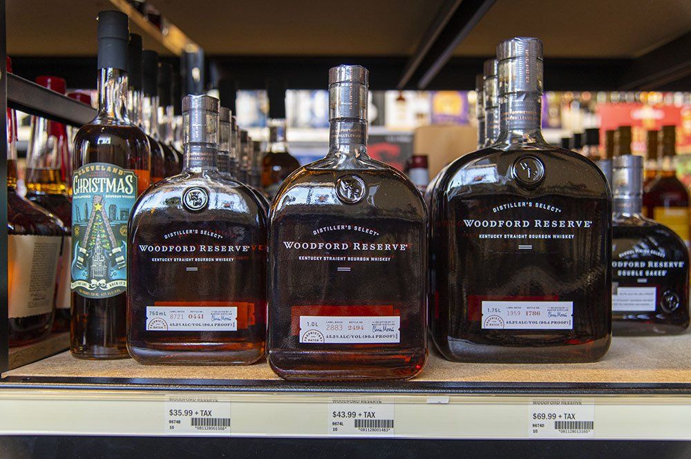 Bottles of brown liquor on a shelf in a store. The bottles are dark, and there are other bottles in the background.