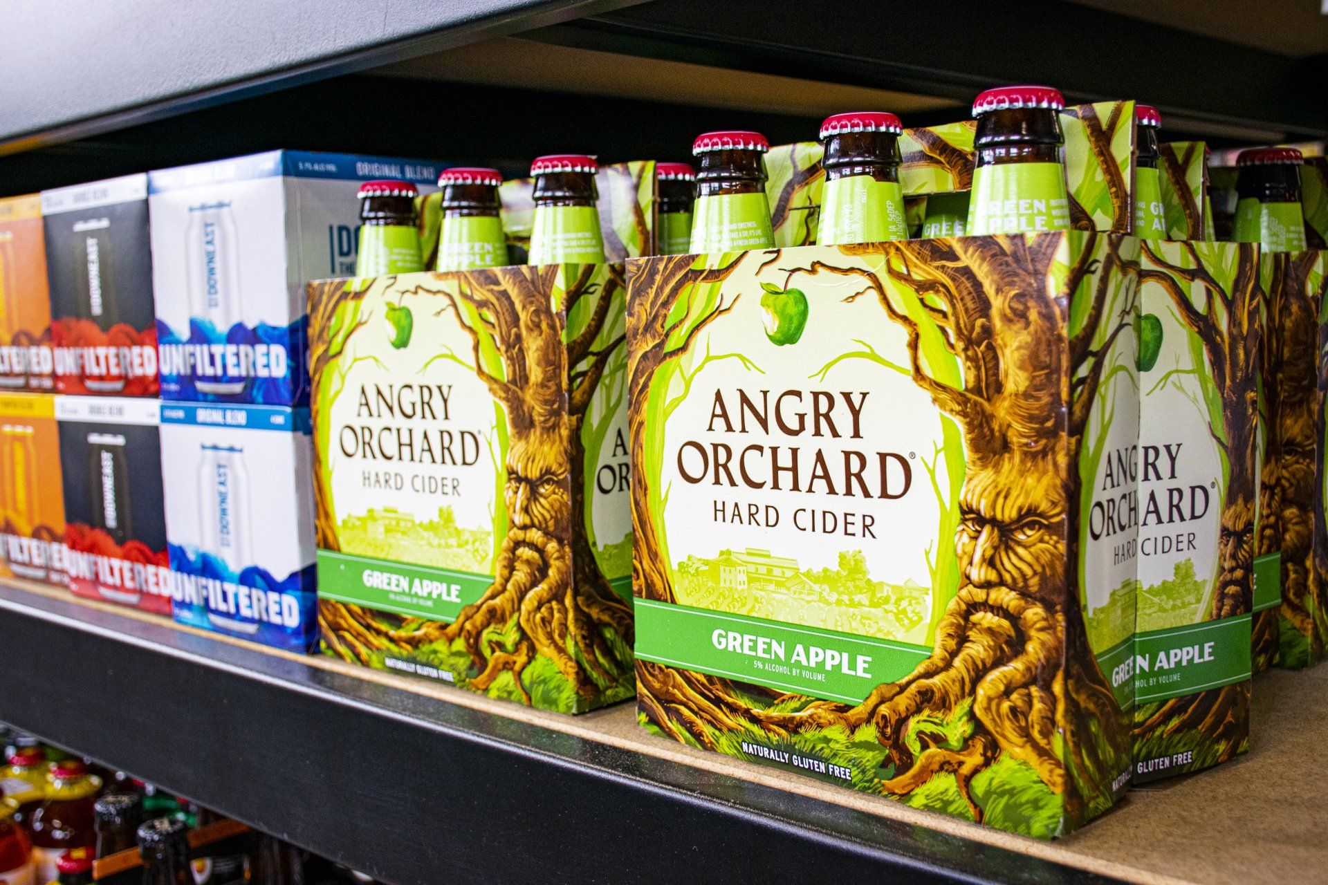 Six-packs of Angry Orchard hard cider on a shelf, in a store. Green and brown packaging.
