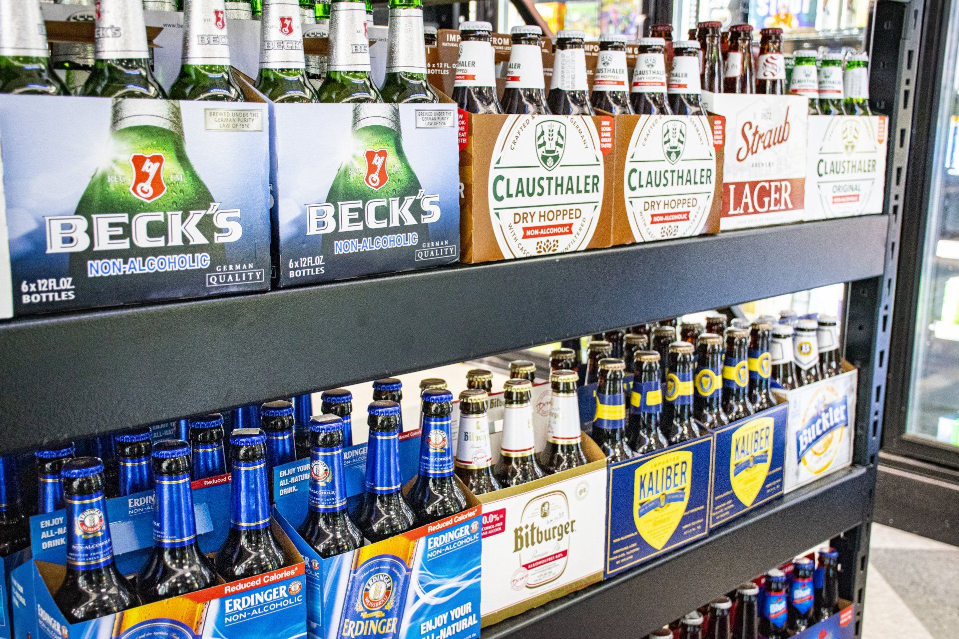 Shelves stocked with various beer brands in six-packs, including Beck's, Clueth, and other brands, in a store setting.