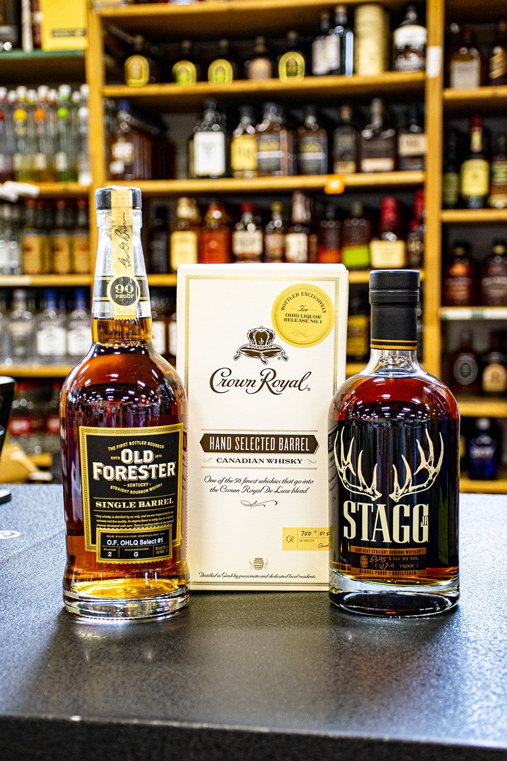Bourbon bottles of Old Forester and Stagg stand next to a white box on a bar, with liquor bottles in the background.