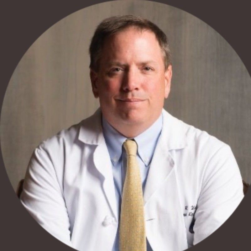 Man in white coat and tie, seated, looking at the viewer.
