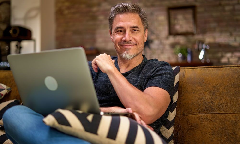 Man sits on couch with laptop, smiling at the camera.
