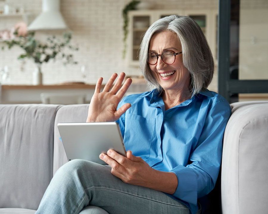 Woman with gray hair waves during a video call on a tablet, smiling, sitting on a couch.