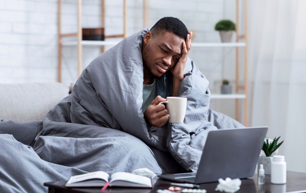 A man wrapped in a blanket is sitting on a couch with a cup of coffee and a laptop.