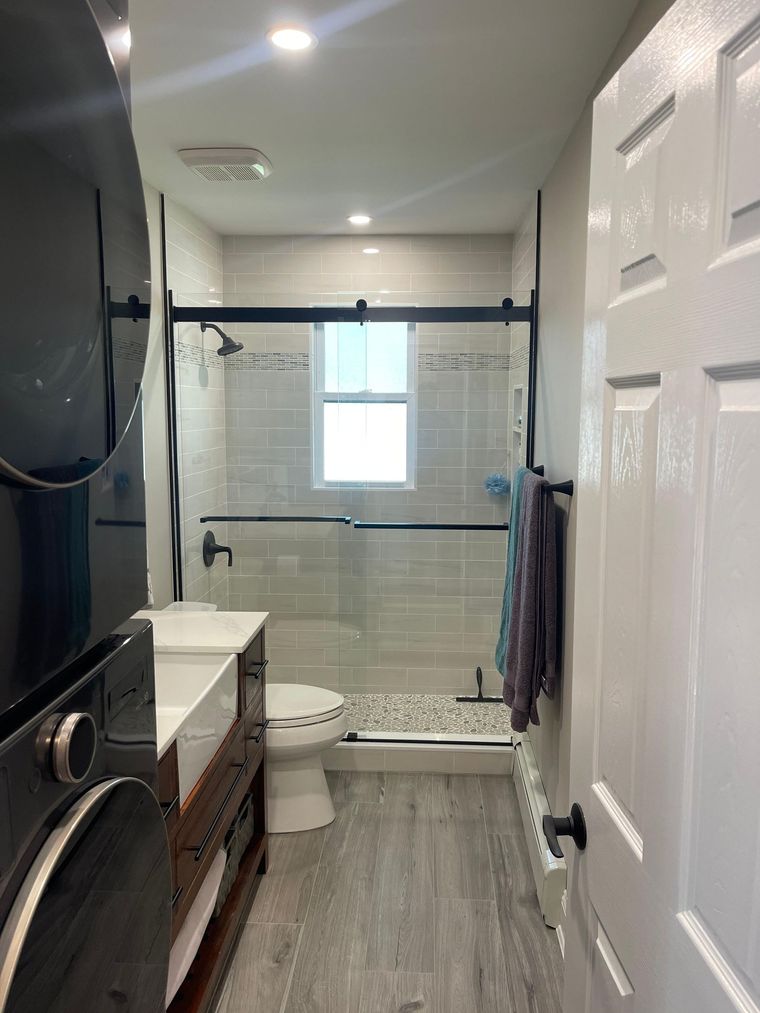 A narrow bathroom featuring a stacked washer-dryer unit, a wooden vanity, a white toilet, and a glass-enclosed shower.