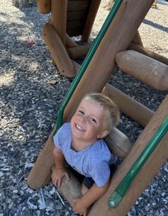Smiling young child in a blue shirt, playing on a wooden playground structure.