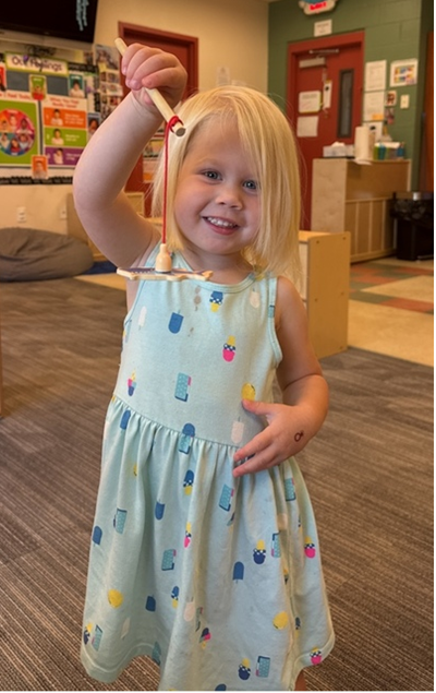 Young girl with blonde hair smiles, holding up a craft in a classroom setting.