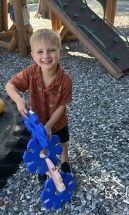 Smiling young boy holding a blue toy at a playground. He's wearing a brown shirt and black shorts.